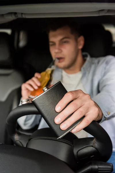 Joven sosteniendo el frasco y bebiendo whisky mientras conduce el coche, fondo borroso - foto de stock