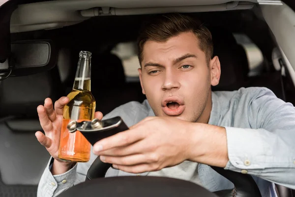 Hombre borracho sosteniendo botella de whisky y frasco mientras está sentado en el coche - foto de stock