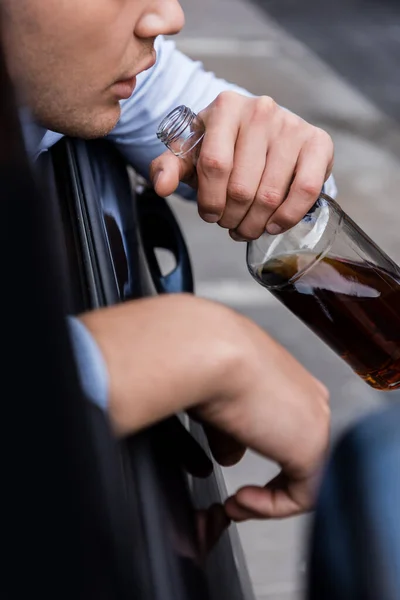 Vista recortada del hombre sosteniendo la botella de whisky en el coche en primer plano borroso, bandera - foto de stock