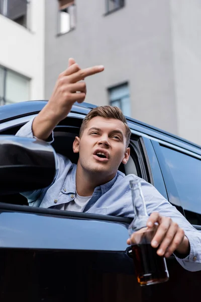 Betrunkener, aggressiver Mann mit Whiskeyflasche zeigt Mittelfinger beim Blick aus dem Autofenster, verschwommener Vordergrund — Stockfoto