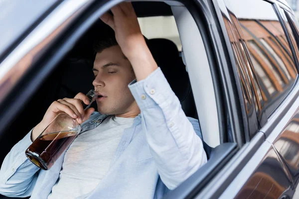 Borracho, somnoliento sentado en el coche y bebiendo whisky con los ojos cerrados - foto de stock