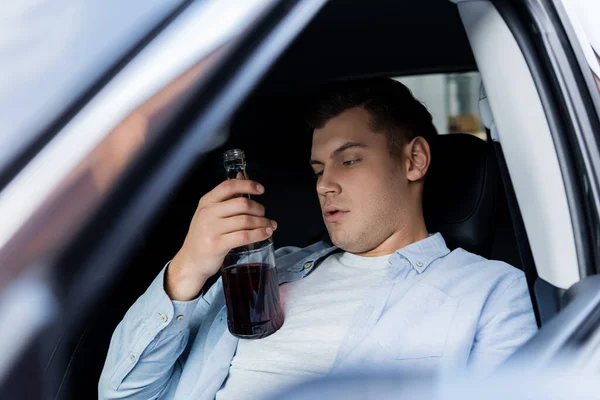 Hombre borracho sosteniendo botella de whisky mientras está sentado en el asiento del conductor en el coche - foto de stock