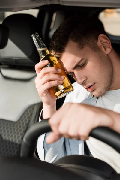 Drunk man holding bottle of alcohol near head while sitting at steering wheel in car, blurred foreground — Stock Photo