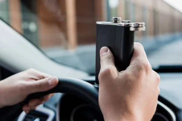 Cropped view of man holding flask with alcohol while driving car, blurred background — Stock Photo