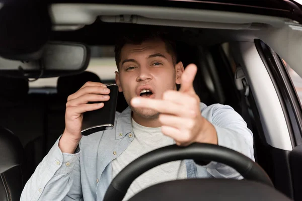 Drunk man holding flask with alcohol and showing middle finger in car, blurred foreground — Stock Photo