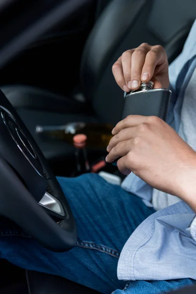 Cropped view of man opening flask with alcohol while sitting in car at drivers seat — Stock Photo