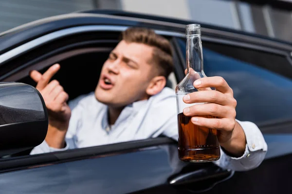Borracho con botella de alcohol mostrando ven aquí gesto mientras mira por la ventana del coche, borrosa primer plano - foto de stock