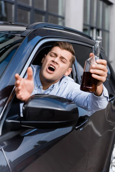 Ubriaco, arrabbiato uomo con bottiglia di alcol gesticolando e urlando guardando fuori dal finestrino dell'auto, in primo piano sfocato — Foto stock