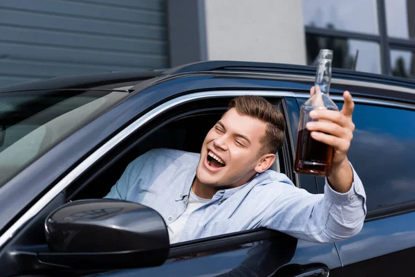 Excitado, borracho hombre con botella de whisky gritando mientras mira por la ventana del coche - foto de stock
