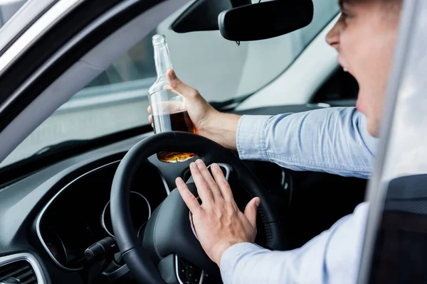 Vista cortada de bêbado, homem agressivo com garrafa de álcool gritando e bip durante a condução do carro, foreground borrado — Fotografia de Stock