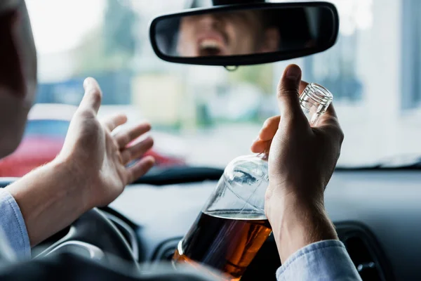 Partial view of angry drunk man with bottle of whiskey shouting and gesturing while driving car, blurred foreground — Stock Photo