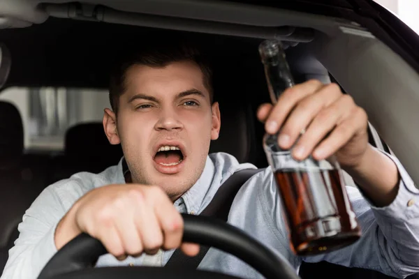 Drunk, aggressive man shouting while driving car and holding bottle of alcohol on blurred foreground — Stock Photo