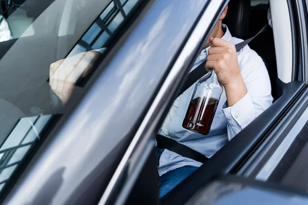 Vista parcial del hombre sosteniendo la botella de whisky mientras conduce el coche, borrosa primer plano - foto de stock