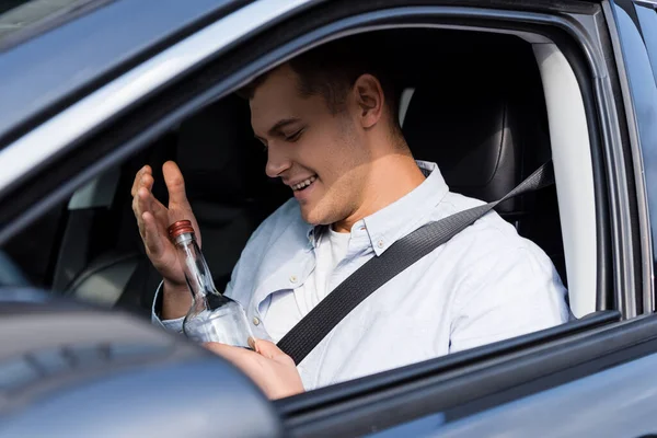 Drunk man smiling and holding bottle of whiskey while sitting in car on blurred foreground — Stock Photo