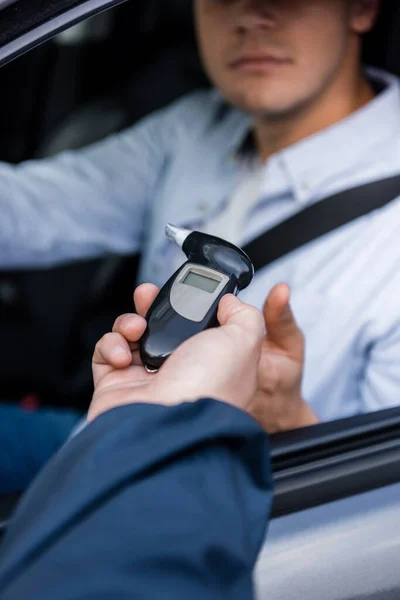 Cropped view of policeman giving breathalyzer to driver sitting in car on blurred background — Stock Photo