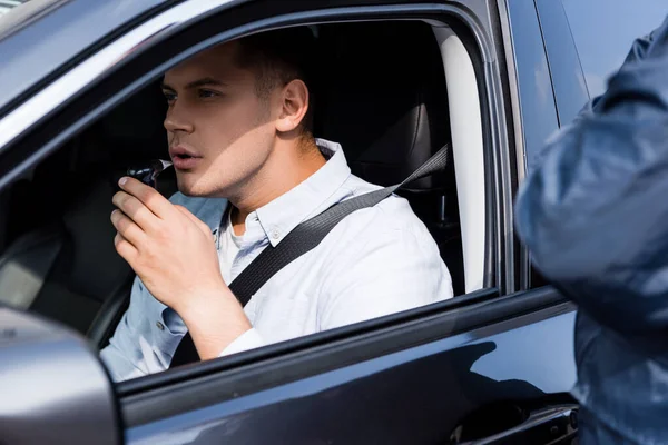 Young man in car blowing into breathalyzer, and policeman standing on blurred foreground — Stock Photo