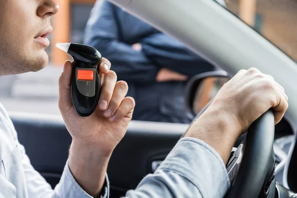 Cropped view of driver blowing into breathalyzer, and policeman standing near car on blurred background — Stock Photo