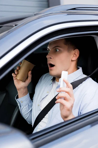 Uomo scioccato che tiene il caffè per andare e il telefono cellulare al posto del conducente in auto, primo piano sfocato — Foto stock