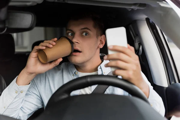 Surprised man drinking coffee to go and using smartphone at steering wheel in car, blurred foreground — Stock Photo