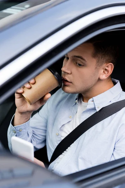 Человек сообщения на мобильном телефоне и пить кофе, чтобы пойти в машине на размытом переднем плане — стоковое фото