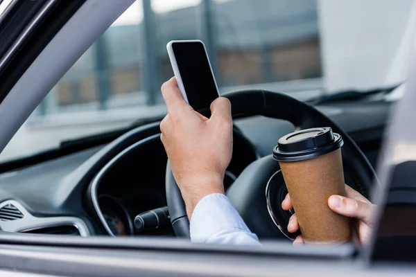 Частковий вид людини, що тримає каву, щоб піти і спілкуватися на смартфоні в машині на розмитому передньому плані — стокове фото