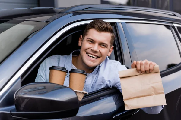 Веселый водитель, держа в руках кофе и бумажный пакет, улыбаясь в камеру — стоковое фото