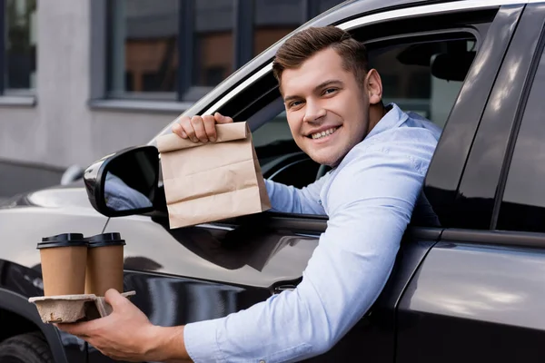 Happy man holding disposable cups and paper bag while sitting in car and looking at camera — Stock Photo