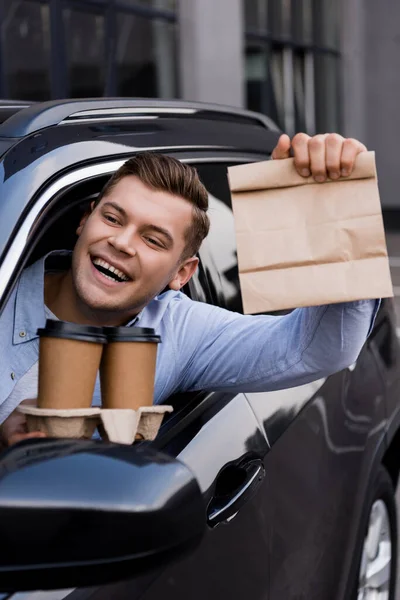 Homem feliz segurando copo descartável e saco de papel enquanto sentado no carro — Fotografia de Stock