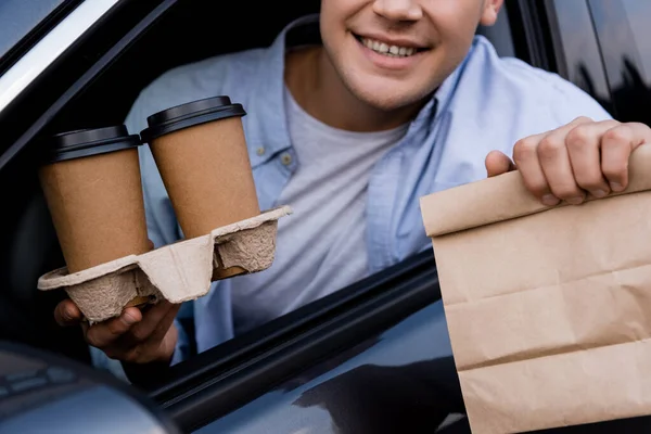 Cropped view of smiling man holding paper bag and coffee to go in car, blurred foreground — Stock Photo