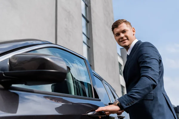Smiling businessman looking at camera while opening car on blurred foreground — Stock Photo