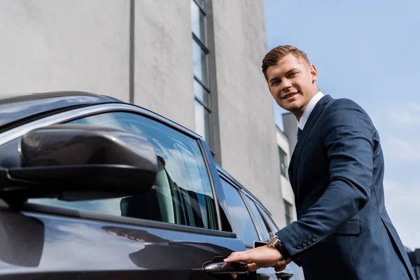 Lächelnder Geschäftsmann blickt in die Kamera, während er sein Auto auf verschwommenem Vordergrund öffnet — Stockfoto