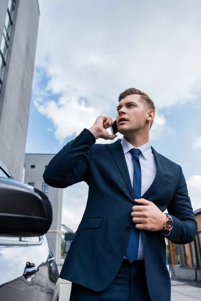 Young businessman unbuttoning blazer against cloudy sky near car on blurred foreground — Stock Photo