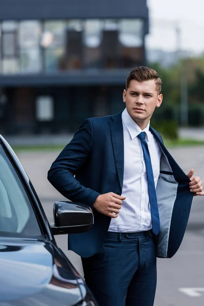 Jeune homme d'affaires décollant blazer près de la voiture au premier plan flou — Photo de stock