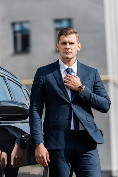 Joven empresario confiado tocando corbata cerca de coche al aire libre - foto de stock