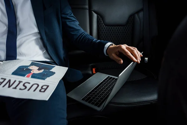 Ausgeschnittene Ansicht des Geschäftsmannes, der Laptop in der Nähe von Zeitung auf dem Rücksitz des Autos berührt — Stockfoto