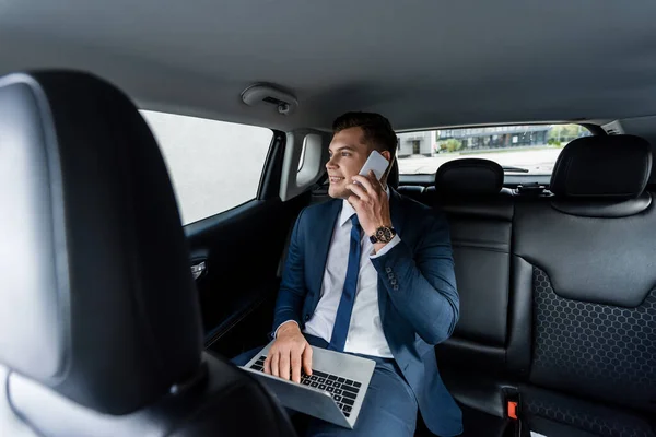 Hombre de negocios sonriente hablando en el teléfono inteligente y utilizando el ordenador portátil en el coche - foto de stock