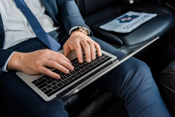 Vista cortada do homem em desgaste formal usando laptop perto de jornal no fundo embaçado no carro — Fotografia de Stock