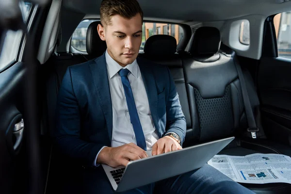 Young businessman using laptop near newspaper on black seat of auto — Stock Photo