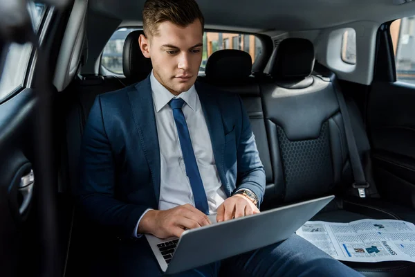 Joven hombre de negocios usando portátil cerca de periódico en asiento negro de auto - foto de stock