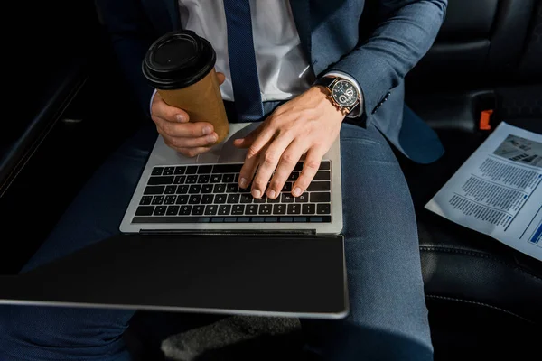 Cropped view of businessman using laptop with blank screen and holding takeaway coffee near newspaper in car — Stock Photo