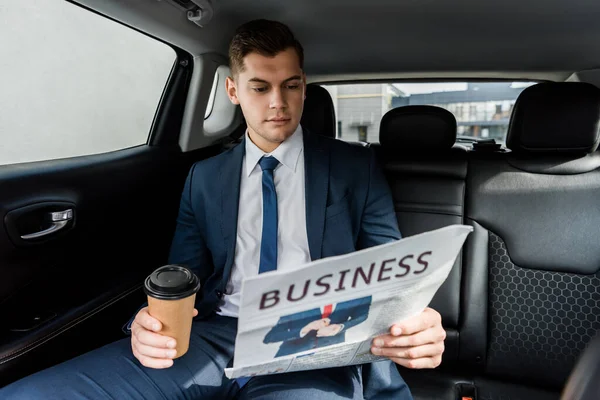 Young businessman holding coffee to go and reading news on blurred foreground in car — Stock Photo