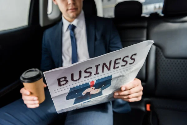 Cropped view of newspaper in hand of businessman with coffee to go in car on blurred background — Stock Photo