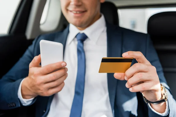 Cropped view of credit card and smartphone in hands of smiling businessman in car on blurred background — Stock Photo