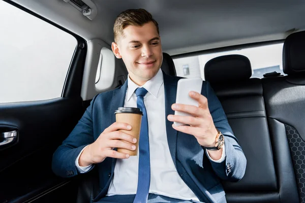 Homme d'affaires souriant utilisant un smartphone et tenant une tasse jetable dans la voiture — Photo de stock