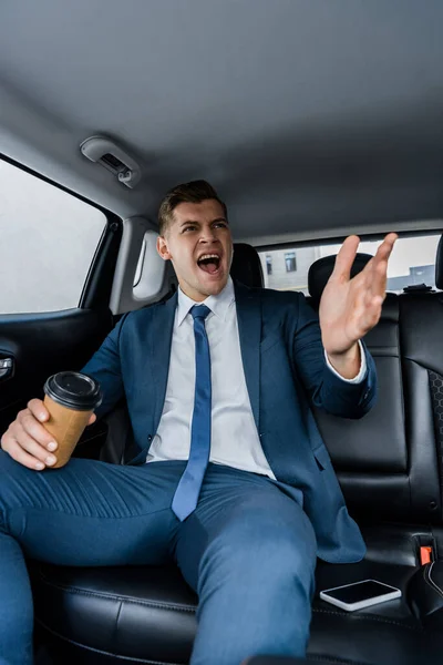Aggressive businessman pointing with hand while holding coffee near smartphone with blank screen in car — Stock Photo