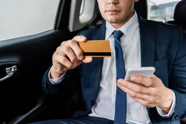 Cropped view of businessman holding credit card and smartphone on back seat of car on blurred background — Stock Photo