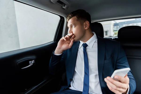 Businessman looking at window and holding smartphone on blurred foreground in car — Stock Photo