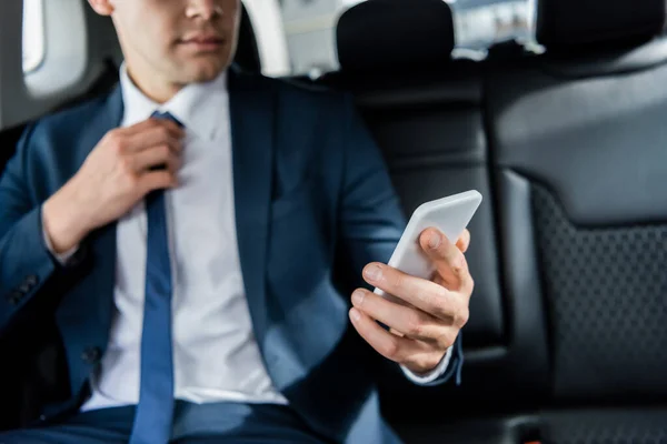 Cropped view of smartphone in hand of businessman adjusting tie on blurred background in car — Stock Photo