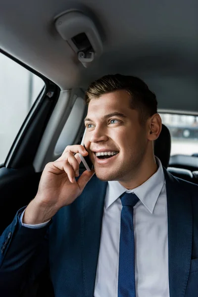 Jovem empresário sorrindo enquanto fala no smartphone no banco de trás do automóvel — Fotografia de Stock
