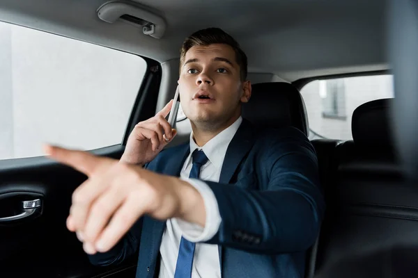 Бізнесмен розмовляє на смартфоні і вказує пальцем на розмитий передній план в машині — стокове фото